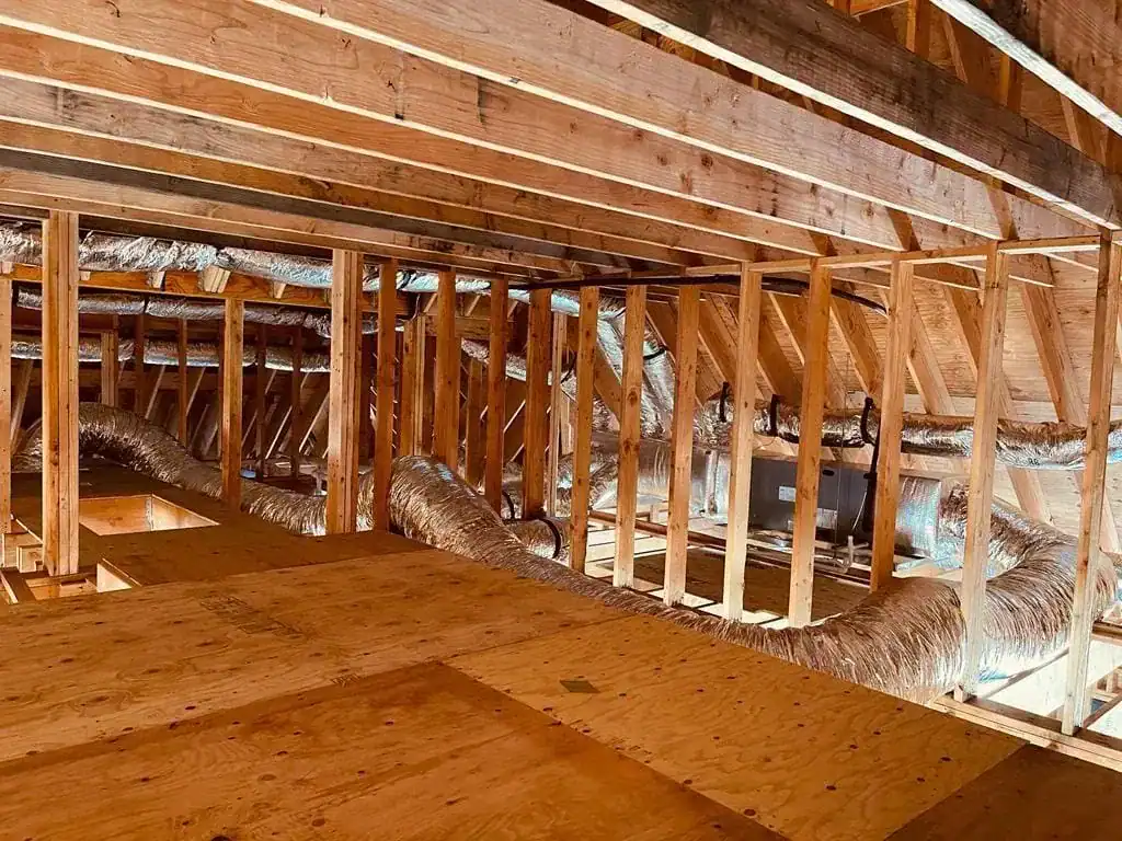 Unfinished attic space with exposed wooden beams, rafters, and plywood flooring. Silver insulated air ducts run through the attic, expertly installed by an HVAC contractor in Nassau County, NY. Sunlight filters in from the right.