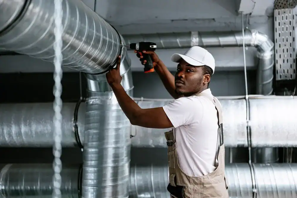 A maintenance worker in overalls and a white cap, employed by an HVAC Contractor Nassau County, uses a power drill to work on large metal air ducts in an industrial NY setting.