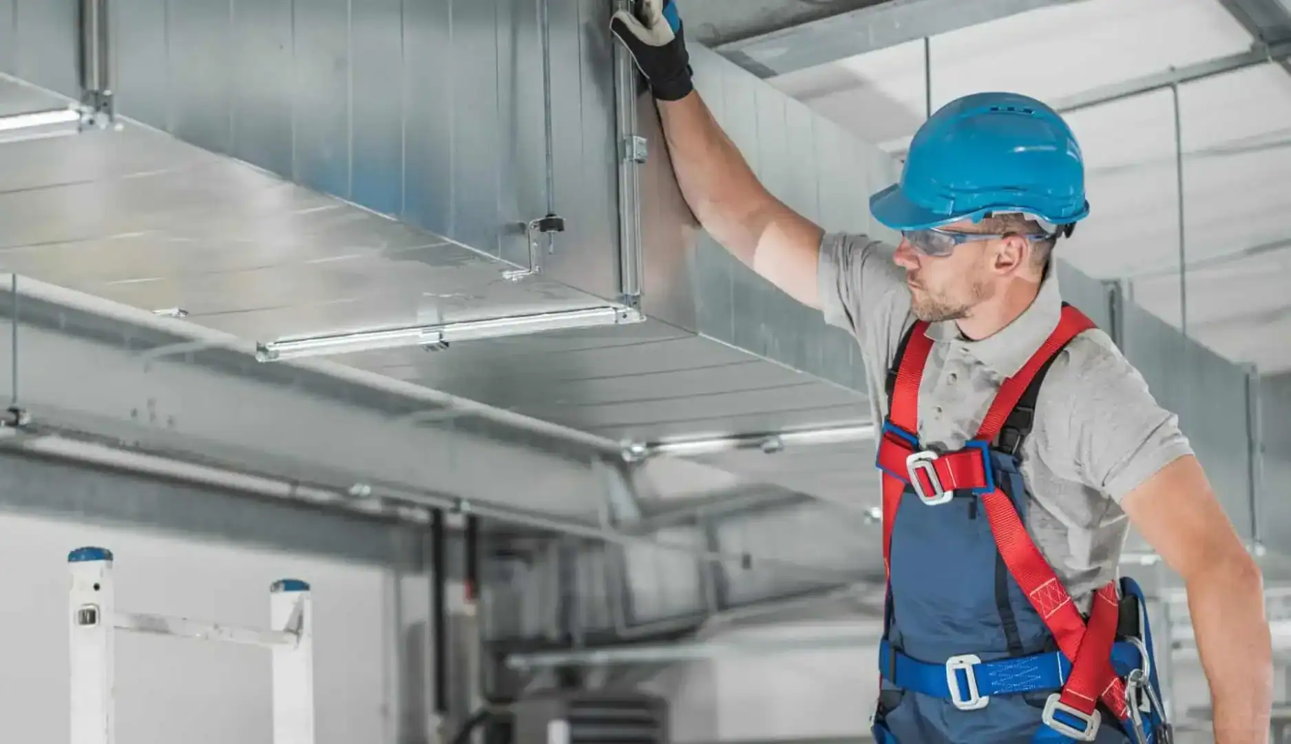 A worker wearing a blue hard hat, safety glasses, and a harness inspects or installs a large metal air ventilation duct for an HVAC Contractor Nassau County in an industrial or commercial building in NY.