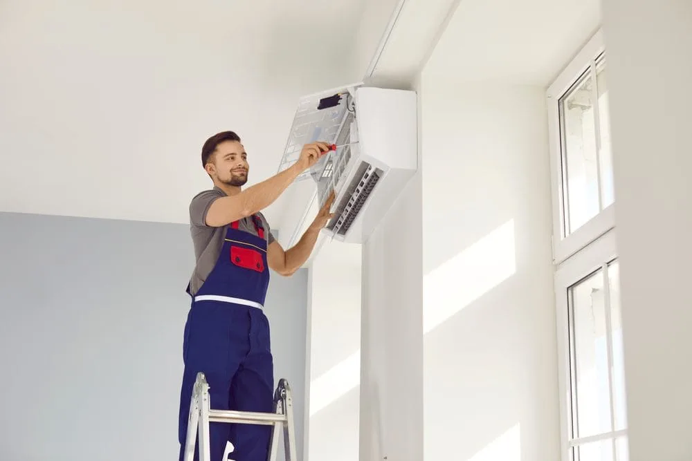 Technician on ladder installing wall-mounted air conditioner in bright room.