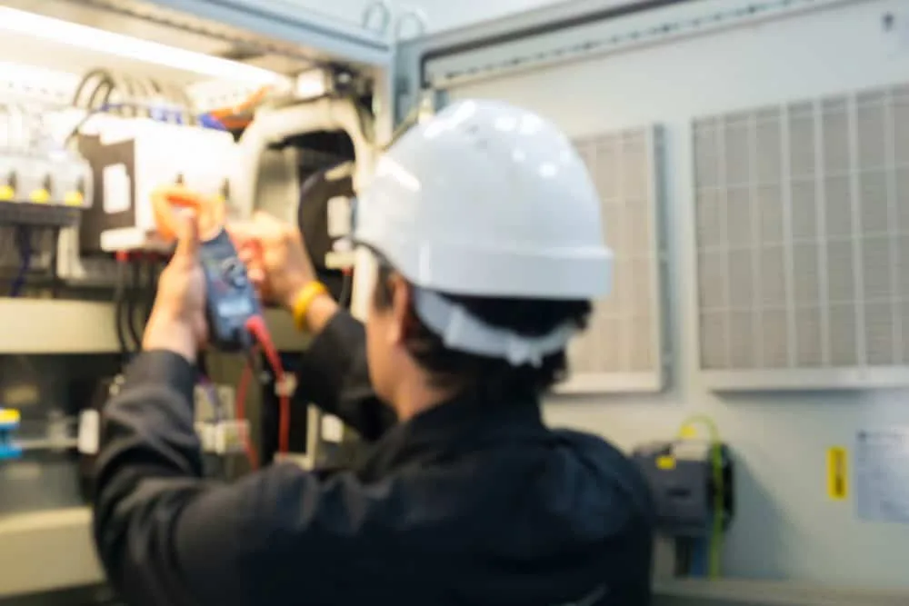 Electrician in hard hat checking control panel wiring with a multimeter.
