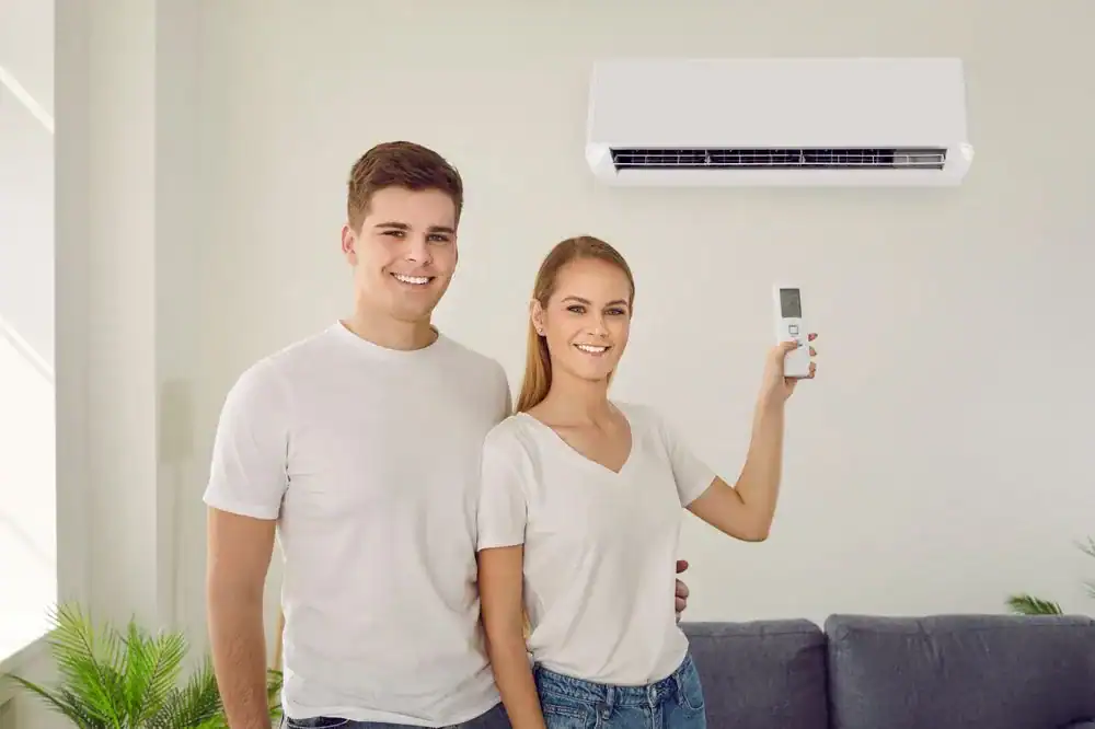 Smiling couple adjusting wall-mounted air conditioner with remote in living room.