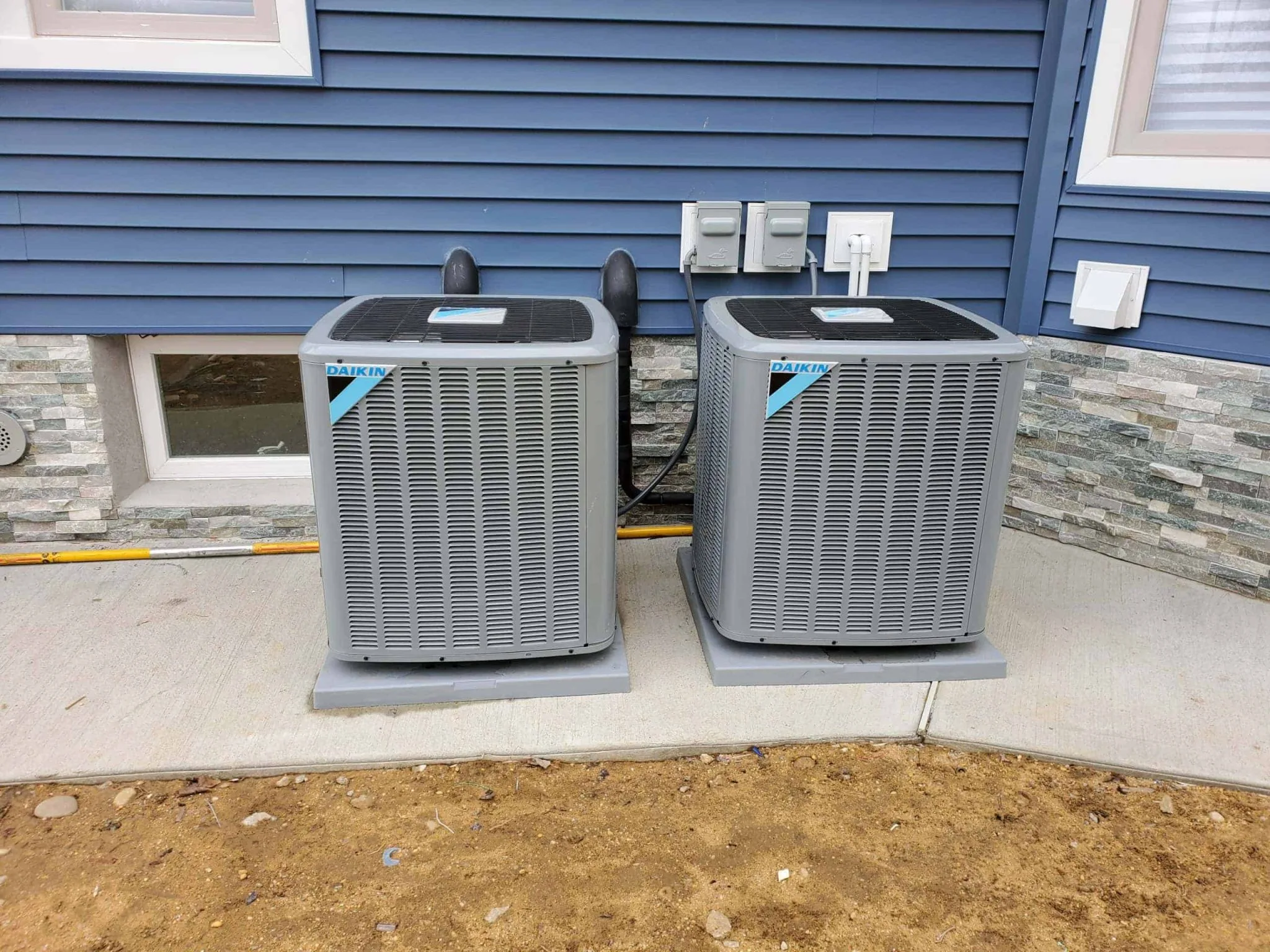Two Daikin air conditioning units next to blue house siding with stone base.