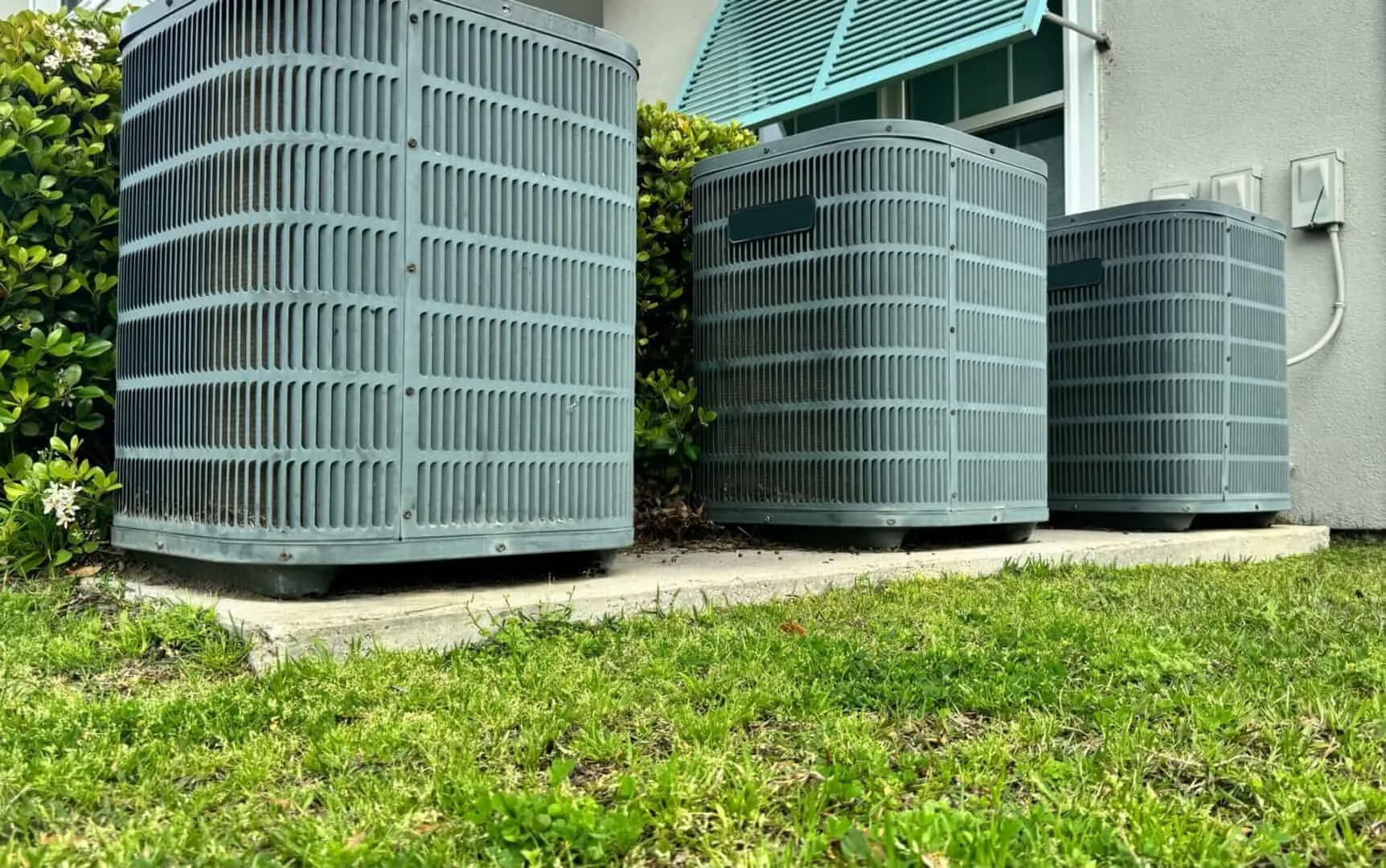 Three outdoor air conditioning units near a house on a concrete slab.