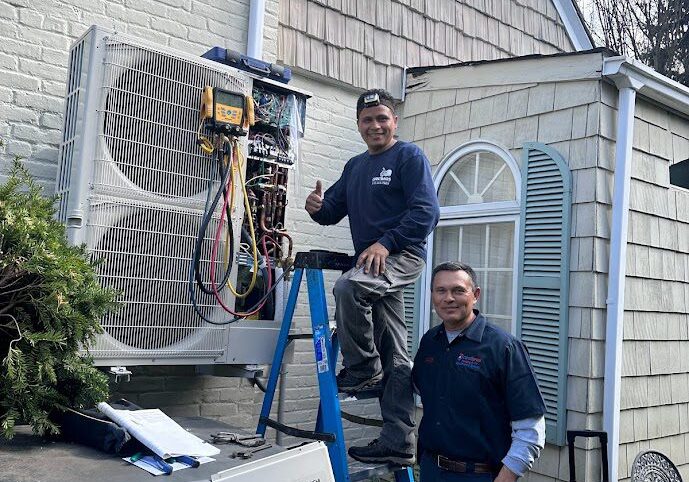 Two technicians working on an outdoor HVAC unit. One is standing on a ladder, giving a thumbs up, while the other stands nearby on the ground. Specializing in Central AC Installation in Nassau County, they're both smiling and wearing work uniforms against the backdrop of shingle siding and a window.